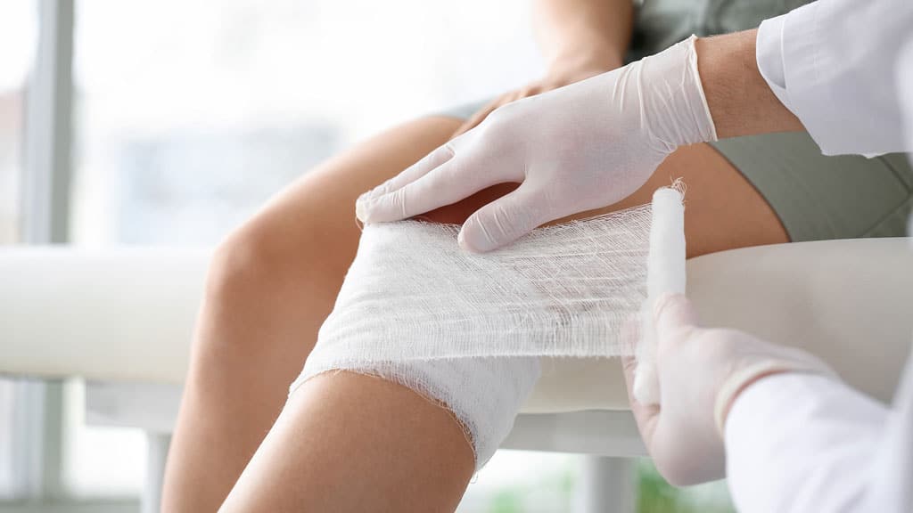 Doctor applying bandage onto knee of young woman in clinic