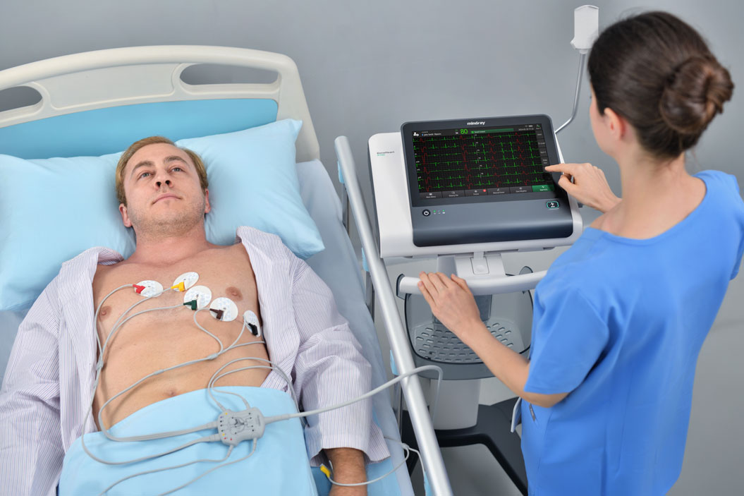 Ein Mann liegt ruhig im Krankenhausbett, mit Elektroden verbunden; eine Frau in blauer Uniform bedient ein Monitoring-Gerät.