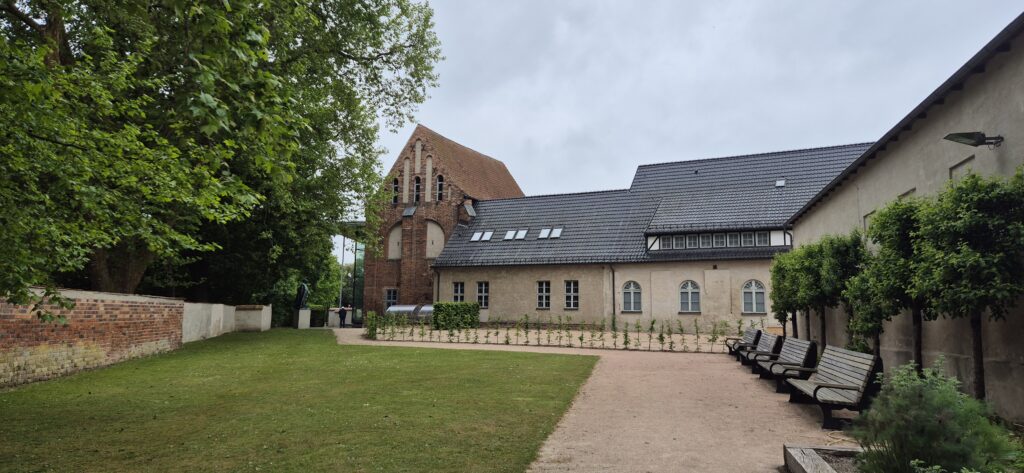 Ein historisches Gebäude mit Ziegeldach, umgeben von Grünflächen und Bänken unter bewölktem Himmel.