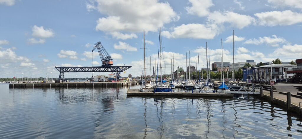 Ein Hafen mit Segelbooten, einem großen Kran und blauem Himmel mit weißen Wolken.